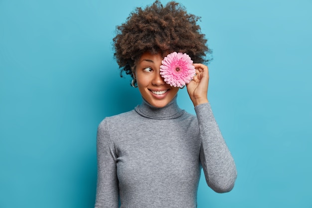 Портрет счастливой энергичной этнической девушки с вьющимися волосами покрывает глаза цветком герберы, позитивно улыбается, носит повседневную водолазку.