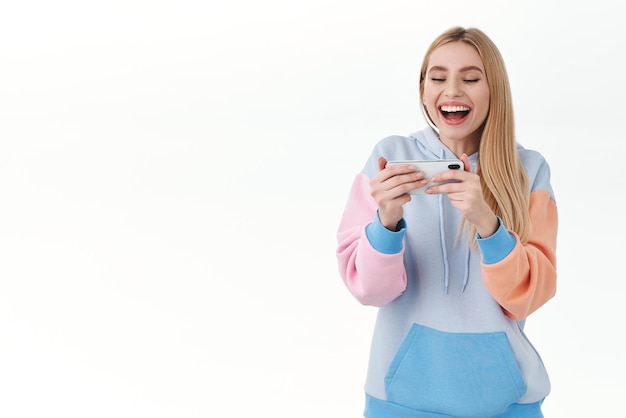 スマートフォンを両手で笑いながら、レースやアーケードの白い背景を再生しているセルの画面を見ているモバイルゲームで勝利を喜んで幸せな熱狂的なブロンドの女の子の肖像画