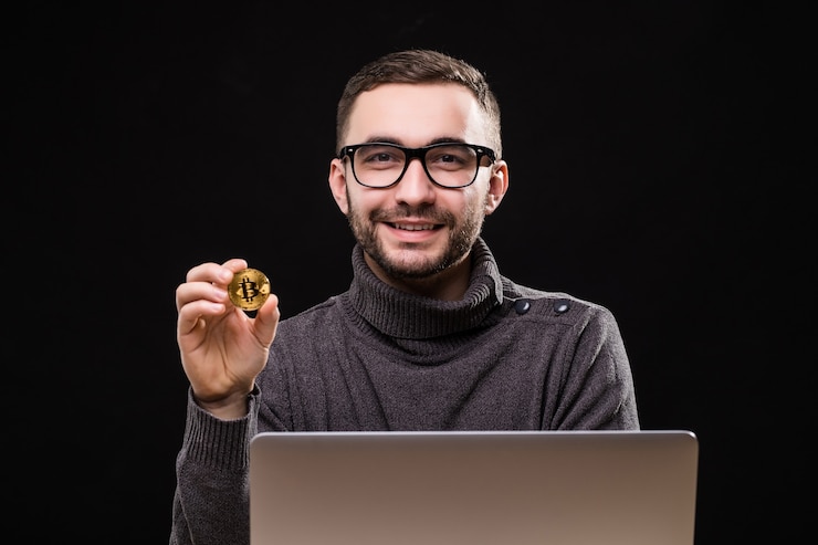 Free Photo portrait of a happy entrepreneur showing bitcoin while sitting at desk with laptop computer isolated over black