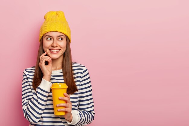 Portrait of happy European woman with glad expression, smiles pleasantly and looks aside, holds takeout coffee, enjoys hot beverage during walk, wears stylish hat and casual jumper, models indoor