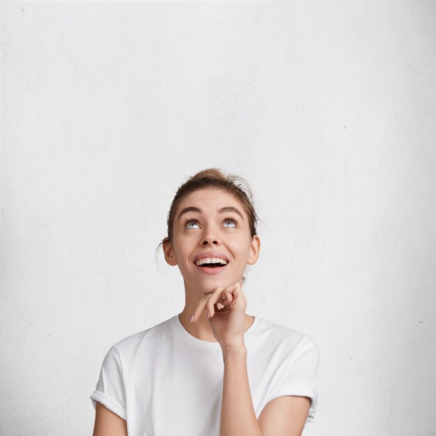 Il ritratto della femmina adorabile felice eccitata felice guarda positivamente verso l'alto, vede qualcosa di piacevole, indossa la maglietta casuale, isolata sopra fondo bianco. persone, felicità e concetto di pubblicità