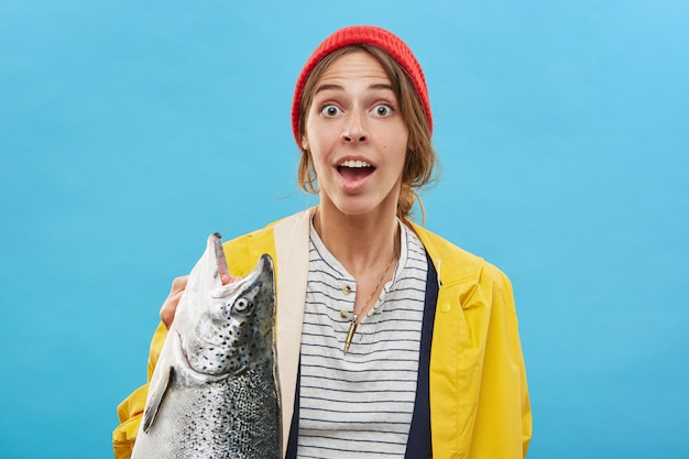 portrait of happy excited young female standing at blank blue wall, holding large freshwater fish, feeling joyful and amazed. People, hobby, activity, leisure and recreation concept