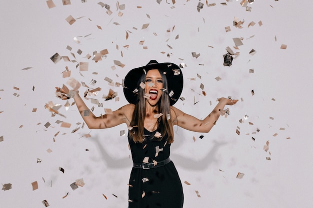 portrait of happy exited woman wearing evening black dress and a hat throwing confetti over isolated wall with true happy emotions.