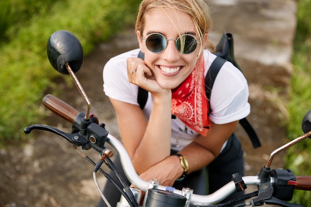 Portrait of happy extremal young woman with shining smile, dressed in fashionable biker`s clothing, rests on fast motorbike, likes her hobby. People, active lifestyle and extreme sport concept