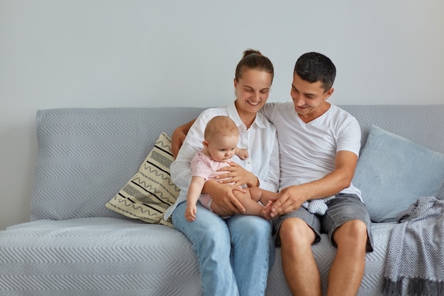 Portrait of happy family sitting on sofa in living room, people wearing casual clothing, spending time with their infant baby at home, parenthood, childhood.