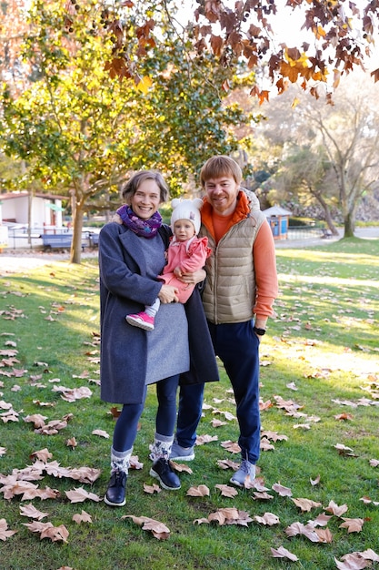 Portrait of happy family standing in autumn park together and smiling. Cheerful mother holding cute baby