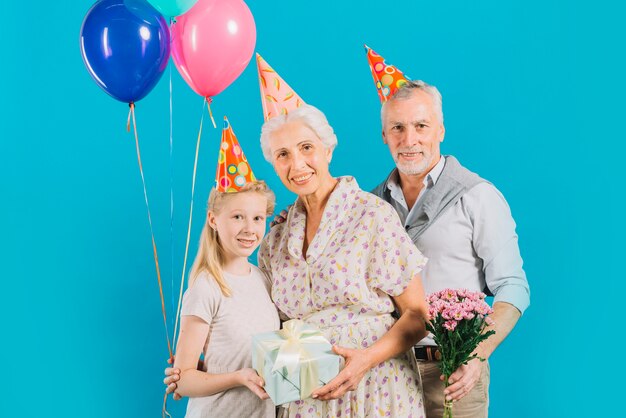 Portrait of happy family with birthday gift; balloons and flowers on blue backdrop