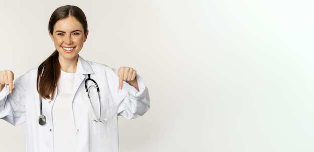 Free photo portrait of happy female doctor pointing fingers down and smiling demonstrating promo offer discount