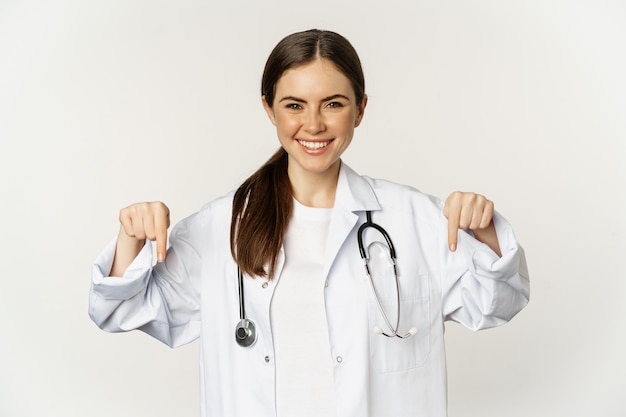 Portrait of happy female doctor pointing fingers down and smiling demonstrating promo offer discount...