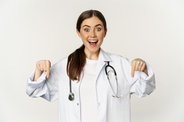 Portrait of happy female doctor pointing fingers down and smiling demonstrating promo offer discount...