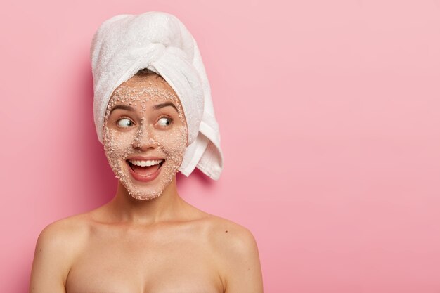 Portrait of happy female model applies sea salt scrub on face, has positive expression, looks aside, has naked body, wears towel after bath