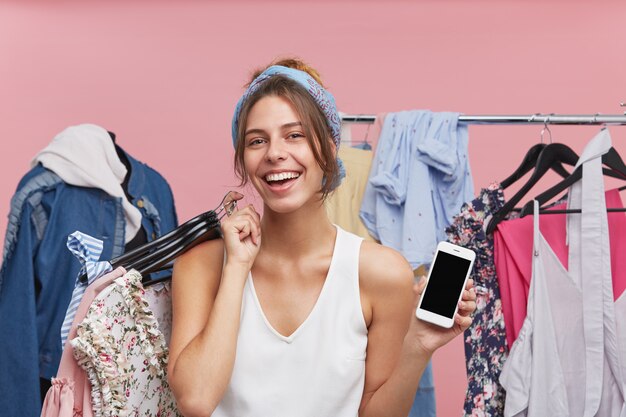 Portrait of happy female wearing white T-shirt, keeping hangers with fashionable clothes and smart phone in hands, having joy while doing shopping, demonstrating blank screen of her mobile phone