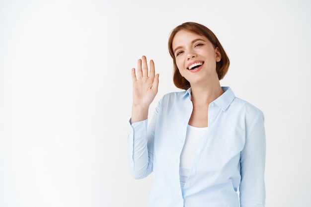 Portrait of happy friendly woman saying hello, waving hand hi greeting gesture, smiling, standing in office blouse on white wall