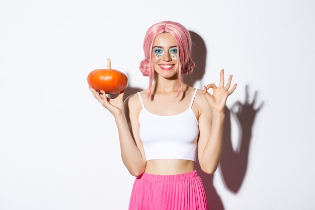 Free photo portrait of happy girl celebrating halloween, showing small pumpkin and okay gesture, wearing cute pink wig.