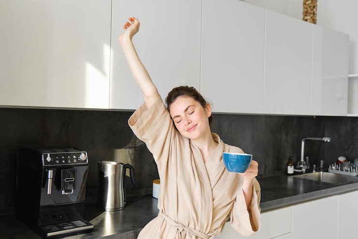 Portrait of happy girl dancing with coffee in the kitchen wearing bathrobe enjoying her morning