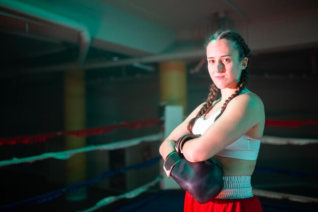 Portrait of happy girl posing on boxing ring. Strong Caucasian girl in sportswear and gloves standing with crossed hands on chest resting and looking at camera. Female boxing and extreme sport concept