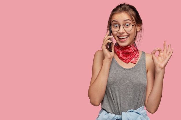 Portrait of happy girl talks on mobile phone with positive expression, makes okay gesture