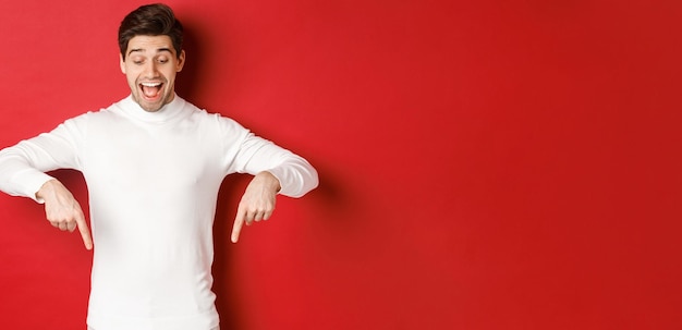 Portrait of happy goodlooking man pointing fingers down and looking at advertisement with excited smile standing over red background