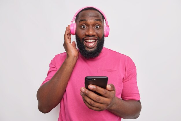 Portrait of happy guy keeps hand on stereo headphones holds mobile phone listens to music wears casual pink t shirt isolated over white wall