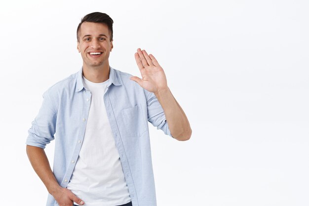 Portrait of happy handsome young man saying hi, waving raised hand informal greeting, nice to meet you or hello sign, smiling pleased, meeting new people joined company, white wall