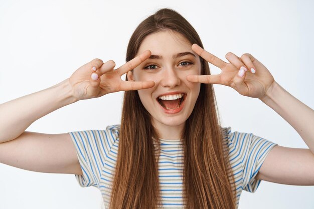 Portrait of happy and healthy girl shows peace, v-sign gesture near eyes and smiling joyful at camera, white background.