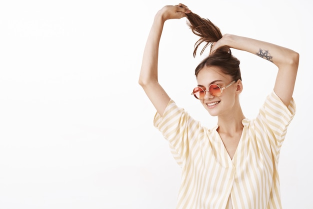 Free photo portrait of happy joyful attractive woman in stylish sunglasses and striped yellow blouse making ponytail