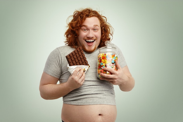 Портрет счастливого и радостного рыжеволосого мужчины в невысокой футболке, стоящего, наслаждаясь нездоровыми, но вкусными сладостями, мармеладом и шоколадом
