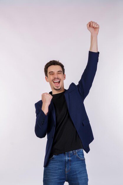 Портрет счастливого радостного молодого бизнесмена, стоящего с жестом победителя, сжимающего кулаки и держащегося изолированным на белом фоне стены студии
