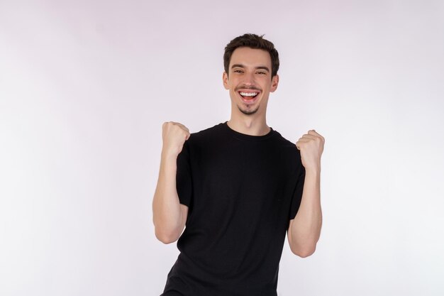 Portrait of happy joyful young man standing doing winner gesture clenching fists keeping isolated on white color wall background studio