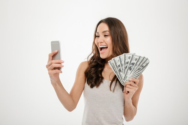 Портрет счастливой радостной женщины 30-х годов, демонстрирующей много денег доллар валюты при использовании своего мобильного телефона, изолированных на белом