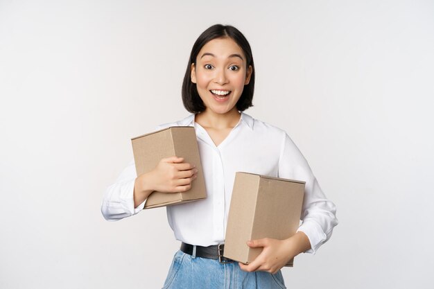 Портрет счастливой корейской девушки, держащей две коробки и улыбающейся, удивленной концепцией камеры шоппинга на белом фоне