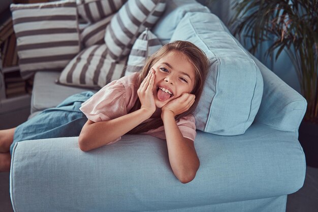 Портрет счастливой маленькой девочки с длинными каштановыми волосами и пронзительным взглядом показывает язык на камеру, лежащей дома на диване.
