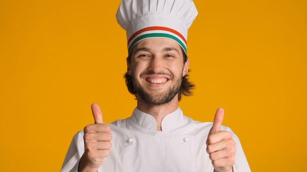 Portrait of happy male chef in uniform keeping thumbs up at camera against a colorful background Handsome man dressed in chef hat showing approved gesture