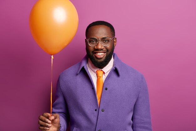 Portrait of happy man comes on stag party, stands with orange balloon, smiles broadly, being in festive mood, congratulates friend