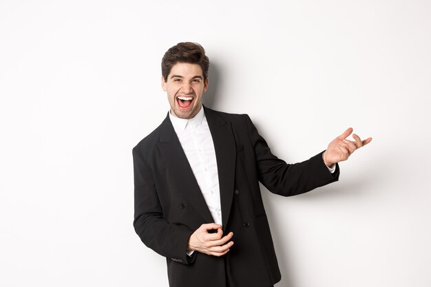 Portrait of happy man dancing at party, playing on invisible guitar and laughing, standing in black suit against white background.