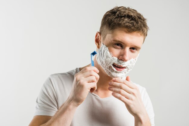 Portrait of happy man looking on camera and shaving his face with razor isolated over gray background