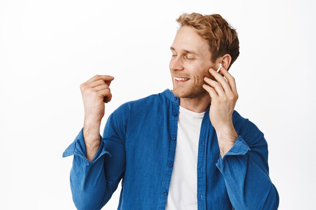 Portrait of happy man smiles and touches headphone while listens to music, snap finger and close eyes, relaxing with favorite songs, stands over white wall