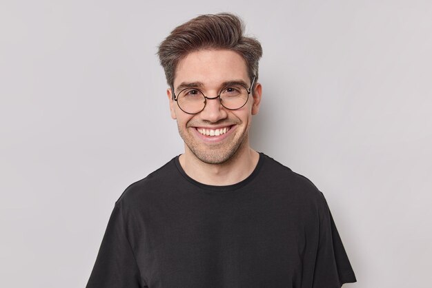 Portrait of happy man with dark hair smiles with white teeth looks confident at camera wears round transparent glasses black t shirt poses at studio against white background. Positive emotions