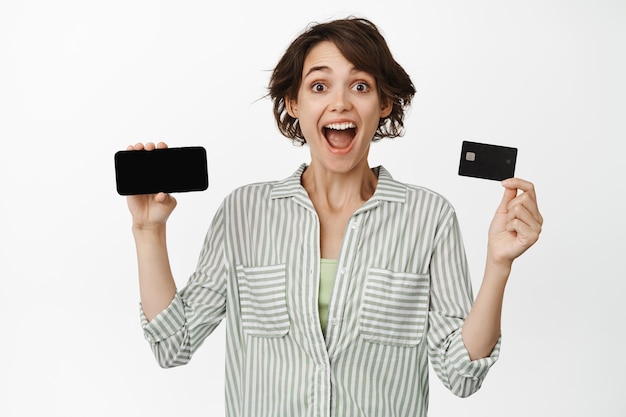 Portrait of happy modern girl shows her credit card, horizontal mobile phone screen, empty display or app interface, standing over white background