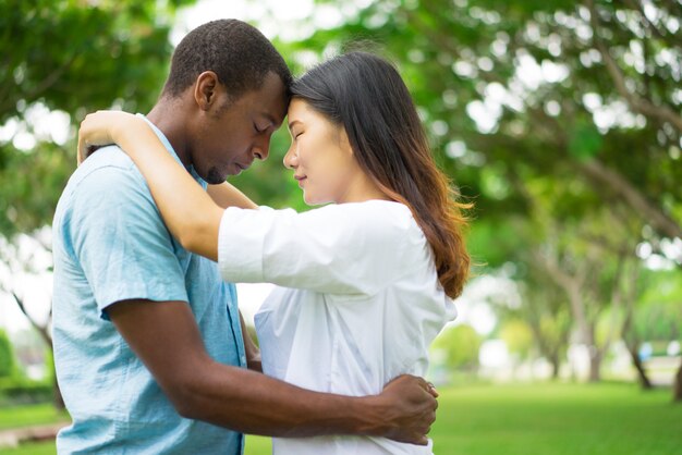 Portrait of happy multiethnic couple standing touching heads in park.