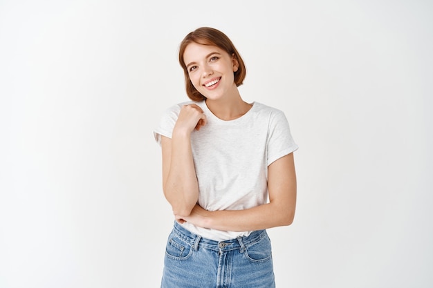 Portrait of happy natural girl, smiling and laughing with joy, looking  carefree, standing in casual clothes against white wall