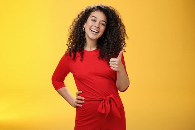 Portrait of happy optimistic young curly-haired woman in red dress laughing joyfully, showing thumbs up in approval and like gesture, delighted with awesome idea, accepting plan over yellow wall.