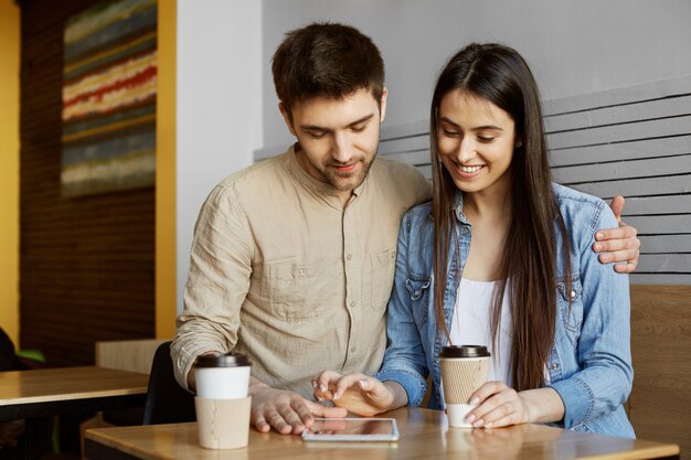 Portrait of happy pair of young people sitting in cafeteria, drinking tea and looking for apartment on digital tablet. Lifestyle concept.