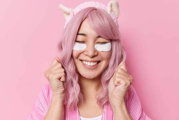 Portrait of happy pink haired woman with eastern appearance clenches fists celebrates special occasion applies white patches for skin refreshment undergoes anti aging procedures wears headband