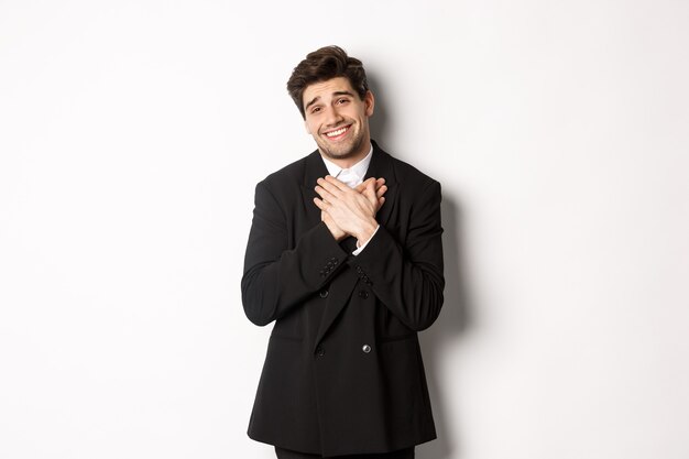 Portrait of happy and pleased handsome man in party suit, holding hands on heart and sighing flattered, saying thank you, standing grateful over white background.