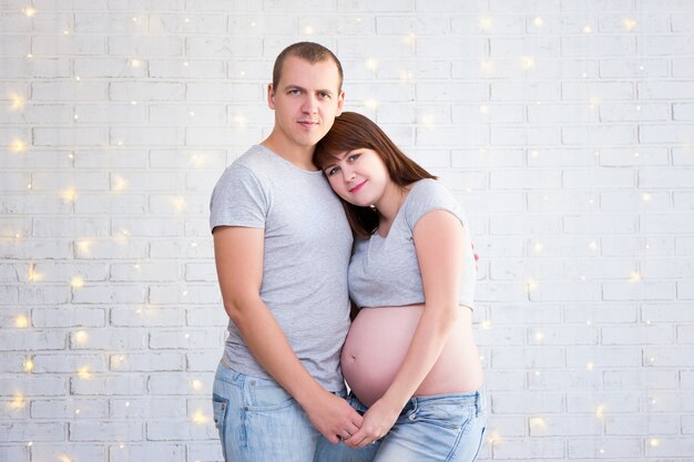 Portrait of happy pregnant couple embracing over white brick wall with christmas lights