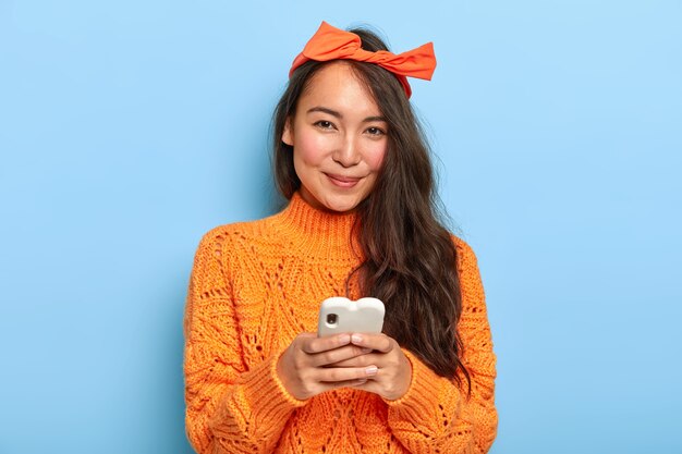 長い髪、ヘッドバンドとオレンジ色のセーターを着て、チャット用の携帯電話を保持し、新しいアプリケーションをダウンロードして幸せなきれいなアジアの女性の肖像画