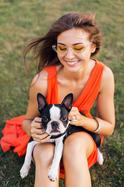 Portrait of happy pretty woman sitting on grass in summer park, holding boston terrier dog, smiling positive mood, wearing orange dress, trendy style, sunglasses, playing with pet, having fun