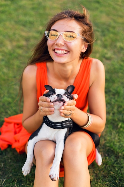 サマーパークの芝生に座って、ボストンテリア犬を抱いて、ポジティブな気分を笑顔、オレンジ色のドレス、流行のスタイル、サングラス、ペットと遊ぶ、楽しんで幸せなきれいな女性の肖像画