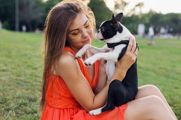 Ritratto di felice bella donna seduta sull'erba nel parco estivo, tenendo il cane boston terrier, sorridente stato d'animo positivo, indossa un abito arancione, stile alla moda, occhiali da sole, giocando con animali domestici
